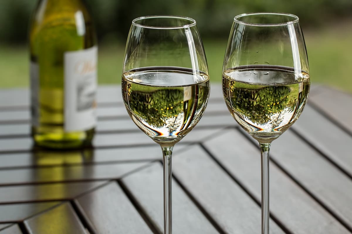 Duas taças de vinho verde em uma mesa de madeira, e ao fundo um garrafa de vinho