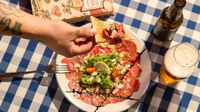 Mesa de bar com prato de carpaccio de carne, rúcula, queijo e limão, acompanhada de cerveja e torrada com aperitivo.