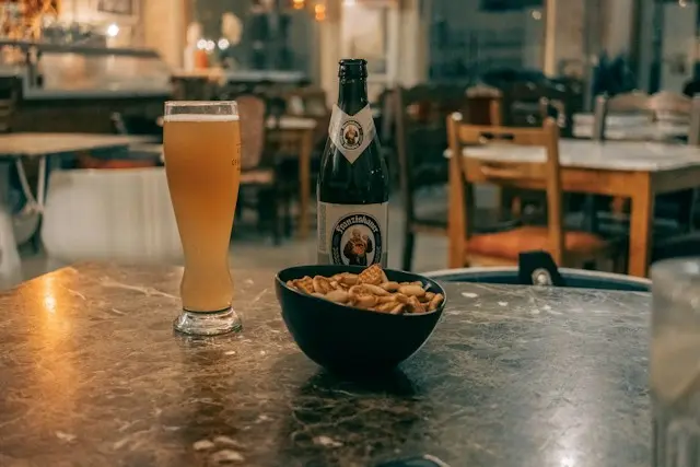 Mesa de bar com copo de cerveja clara, garrafa de cerveja e tigela de amendoins servidos como tira-gosto em ambiente de restaurante.