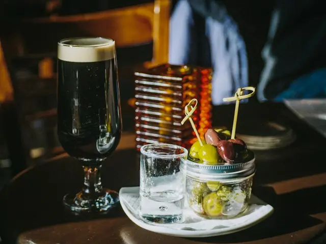 Mesa de bar com taça de cerveja escura, pote de azeitonas verdes e pretas servidas como tira-gosto e um copo pequeno.
