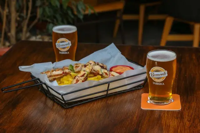 Mesa de bar com dois copos de cerveja clara e porção de frango grelhado com batatas fritas e molho, servida em uma bandeja.