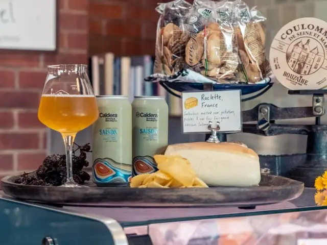Mesa de petiscos com taça de cerveja, duas latas de cerveja artesanal, queijo, batatas chips e acompanhamentos em uma loja de produtos gourmet.