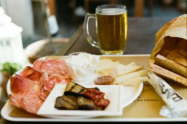 Mesa de bar com tábua de frios, salames, presunto cru, queijos, antepastos, pães e caneca de cerveja clara.