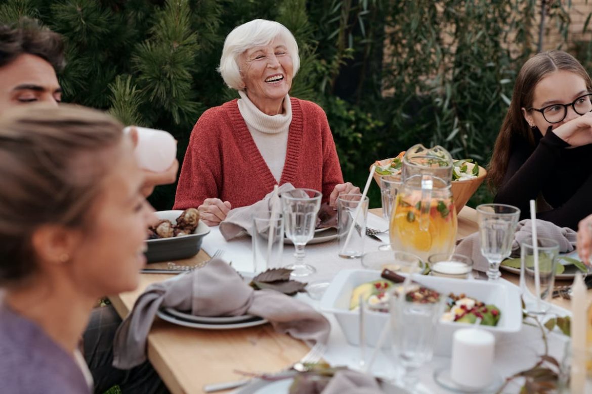 Família reunida para o dia das mães. Na ponta da mesa está uma senhora de cabelos brancos sorridente.
