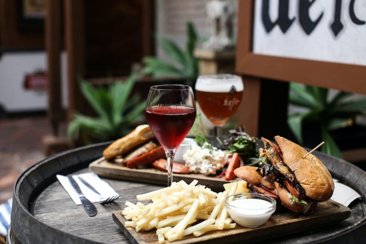 Em uma mesa há um prato com um hambúrguer, batatas fritas e molho, acompanhado de uma taça de vinho
