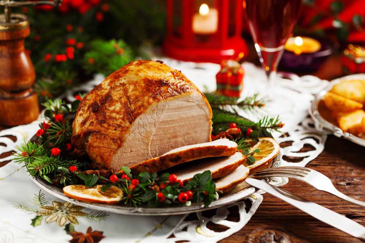 Imagem de um pedaço de pernil assado em um prato decorado com ramos verdes e frutinhas vermelhas. Ao fundo dá para ver uma decoração de natal.