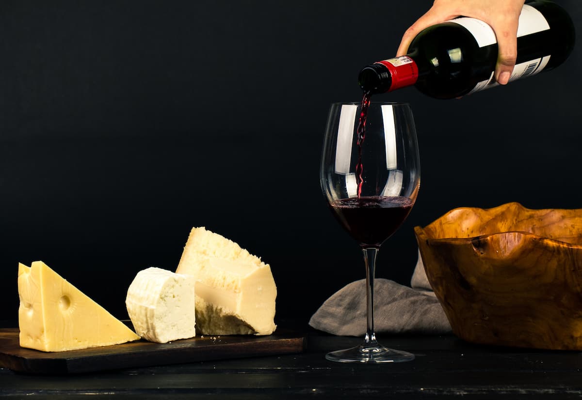Uma pessoa servindo uma taça de vinho com queijos ao fundo