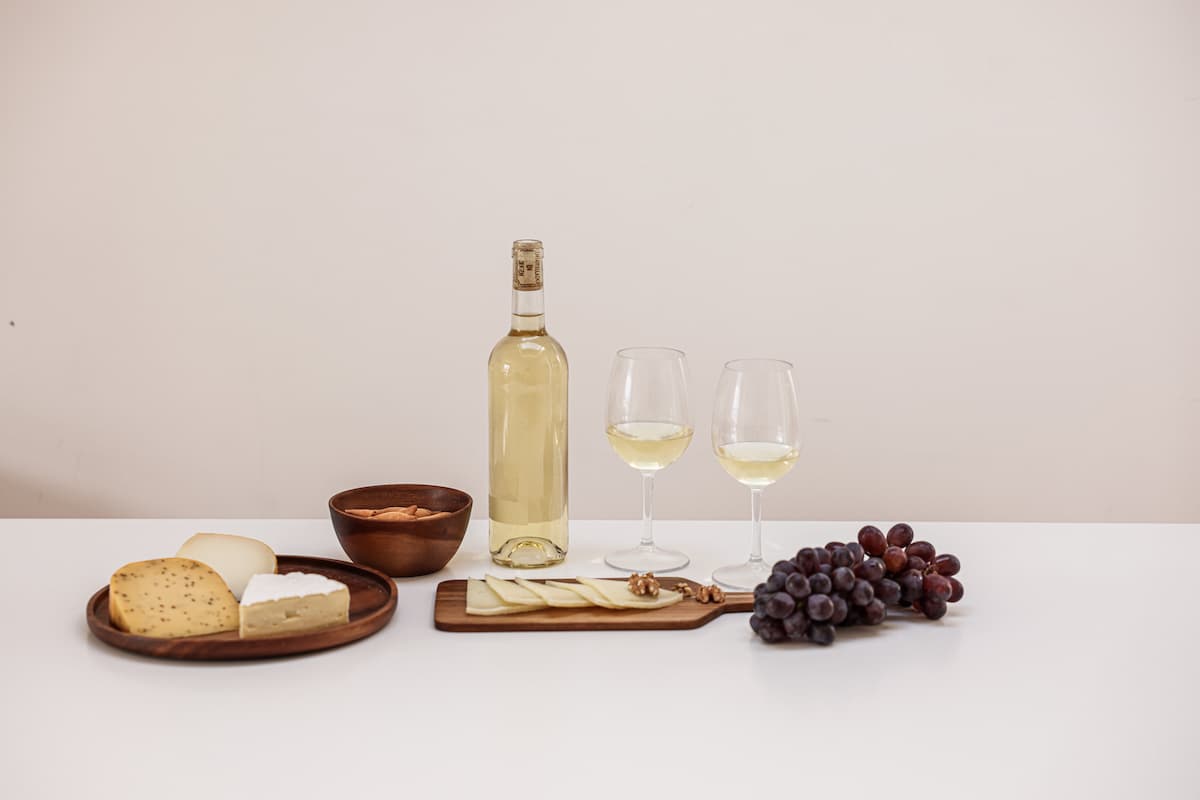 Em uma mesa tem, uma garrafa de champagne, duas taças, uma tábua de madeira com queijo e ao lado, uvas