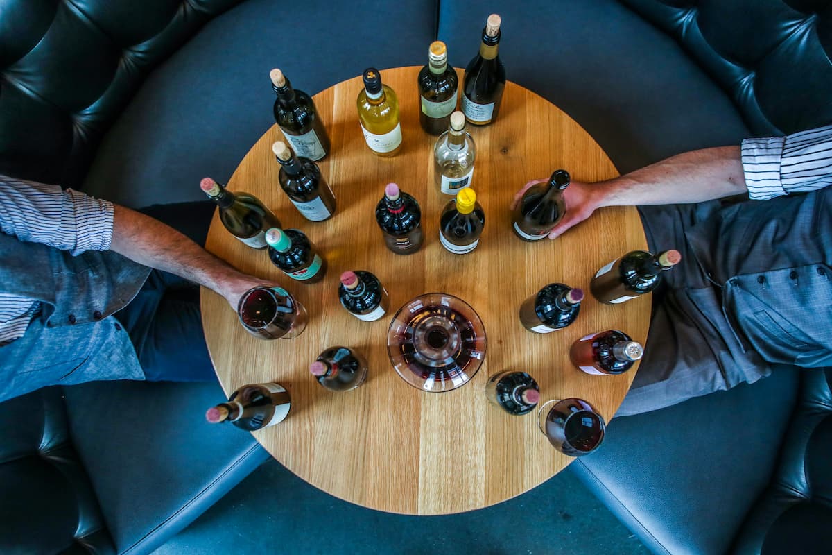 Duas pessoas experimentando diferentes estilos de vinho, e na frente deles, uma mesa cheia de garrafa de vinho