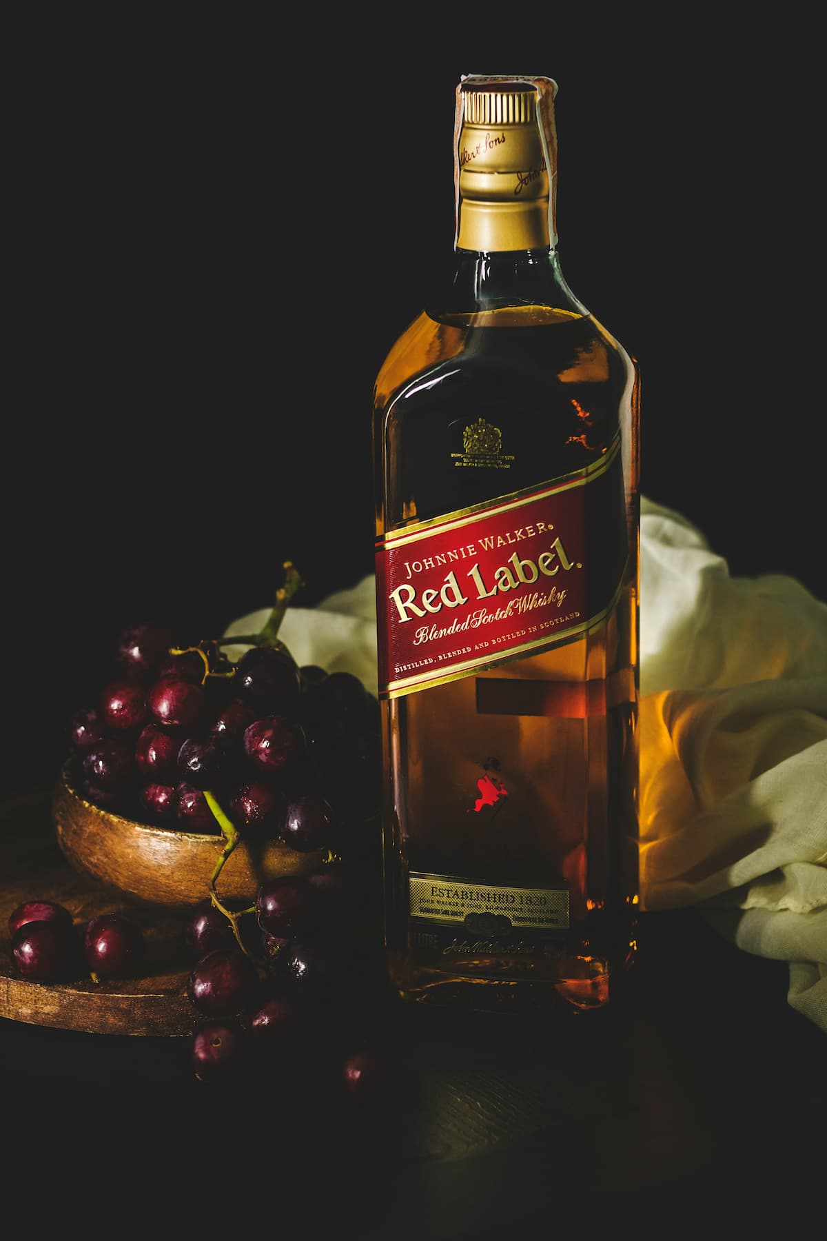 Uma garrafa do Whisky Red Label, em um fundo preto e com um cacho de uva ao fundo