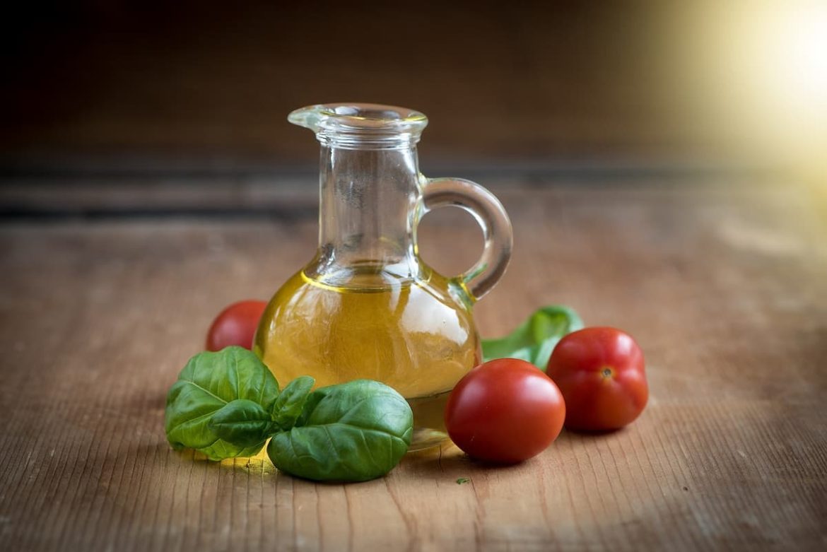 Uma jarra de vidro com azeite dentro, e em volta, dois tomates e duas folhas de hortelã