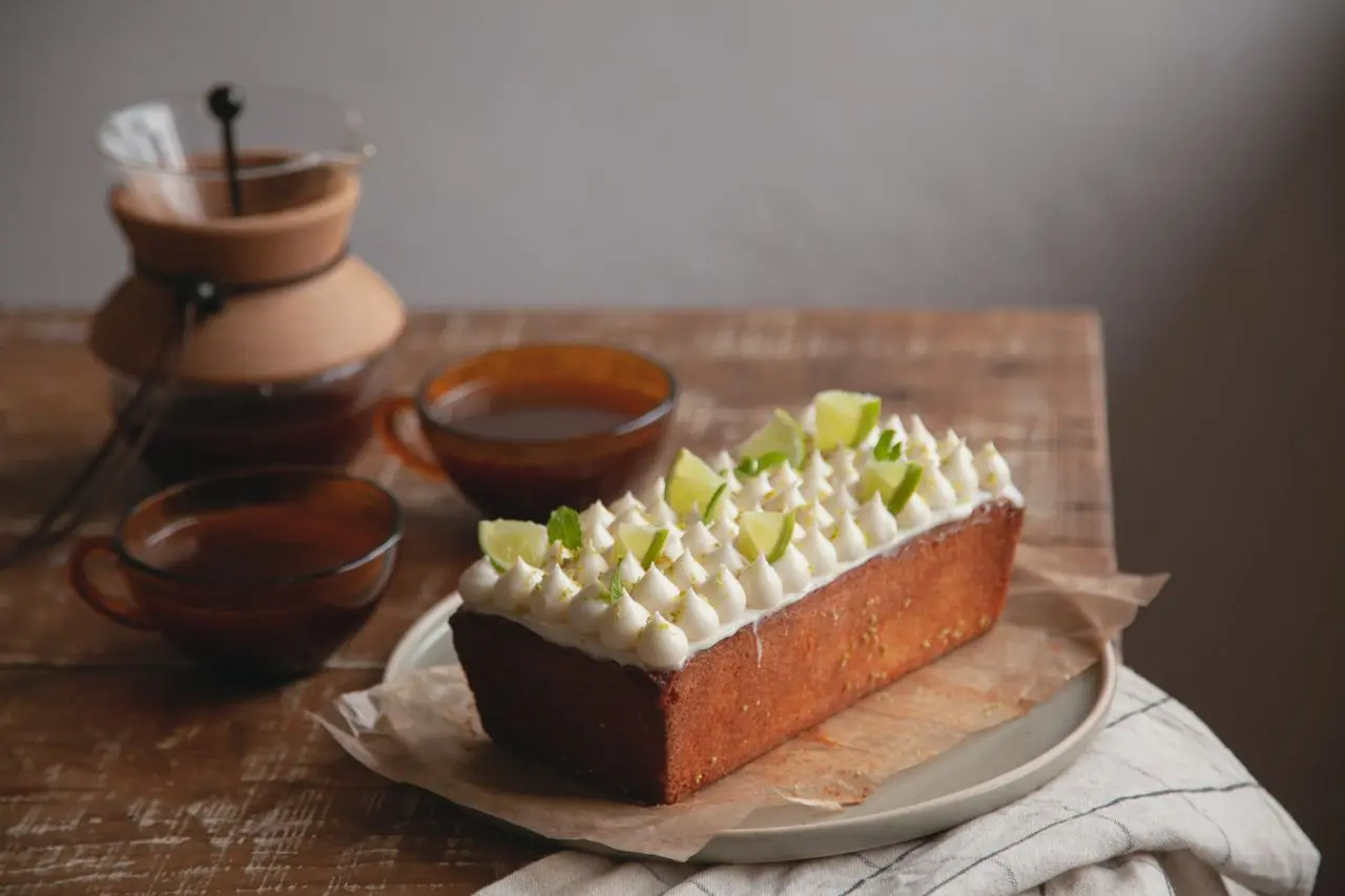 Bolo de limão coberto com creme e pedaços da fruta, servido ao lado de xícaras e cafeteira.
