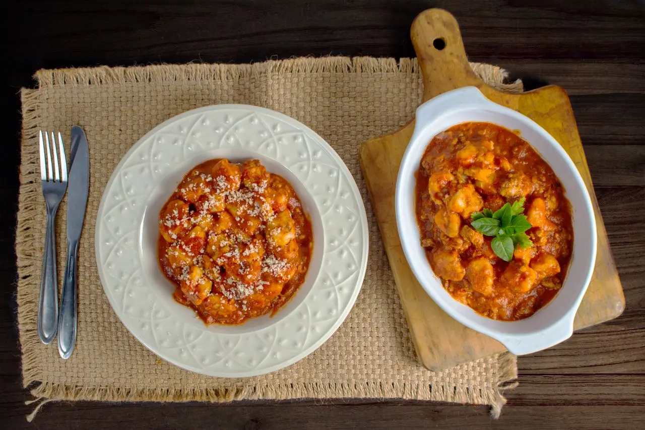 Nhoque ao molho de tomate e queijo ralado, servido em prato e travessa sobre mesa de madeira.