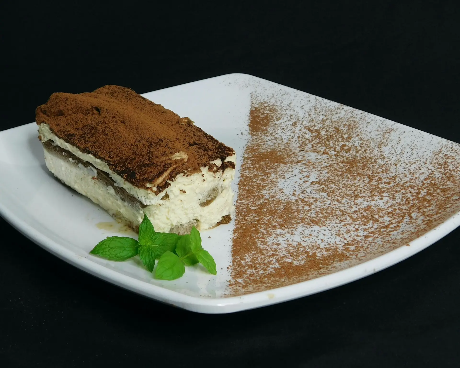 Fatia de tiramisù polvilhada com cacau, servida em prato branco e decorada com folhas de hortelã.