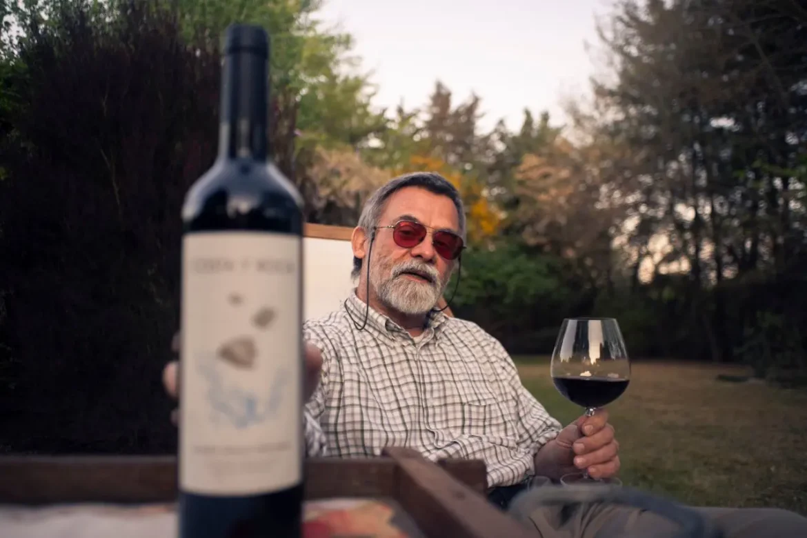 Homem segurando uma taça de vinho tinto ao ar livre, com uma garrafa de vinho em primeiro plano.