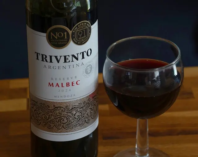 Garrafa de vinho Trivento Reserve Malbec 2024 ao lado de uma taça com vinho tinto sobre mesa de madeira.