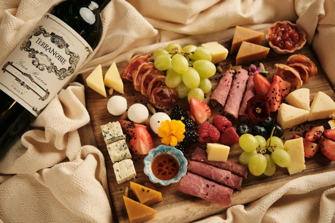 Tábua de frios com queijos, embutidos, uvas, morangos, frutas vermelhas e acompanhamentos, ao lado de uma garrafa de vinho.