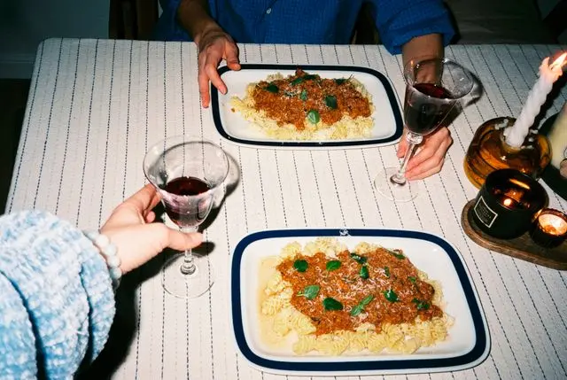 Duas pessoas segurando taças de vinho tinto à mesa, com pratos de macarrão ao molho bolonhesa e velas acesas.