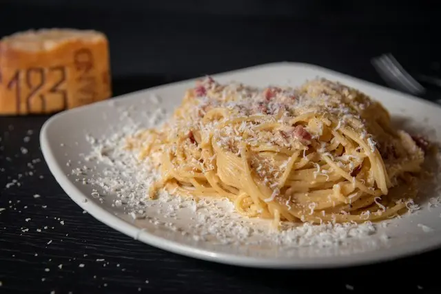 Prato de espaguete à carbonara com queijo ralado e pedaços de bacon, servido em prato branco sobre mesa escura.