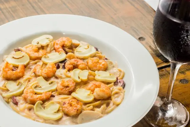 Prato de macarrão com molho cremoso, camarões e cogumelos fatiados, acompanhado de uma taça de vinho tinto sobre mesa de madeira.