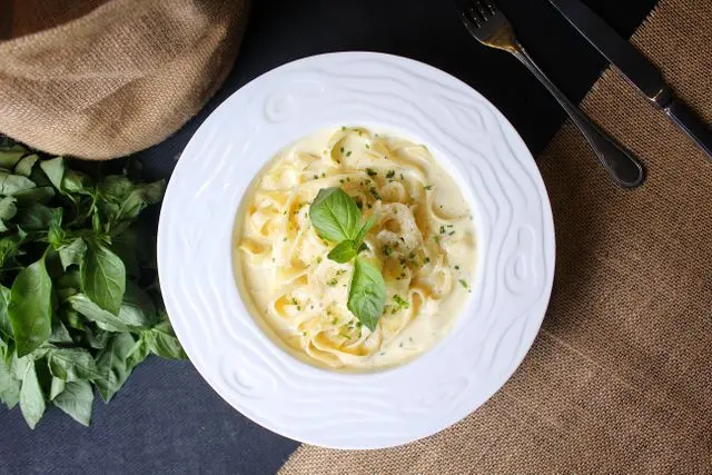 Prato de fettuccine ao molho branco cremoso, decorado com folhas de manjericão e ervas frescas, servido em prato branco.