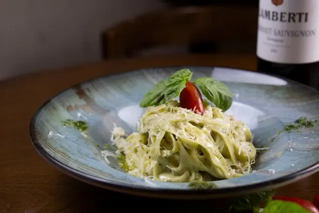 Prato de massa ao molho pesto com queijo ralado, manjericão e tomate-cereja, acompanhado de uma garrafa de vinho Cabernet Sauvignon ao fundo.
